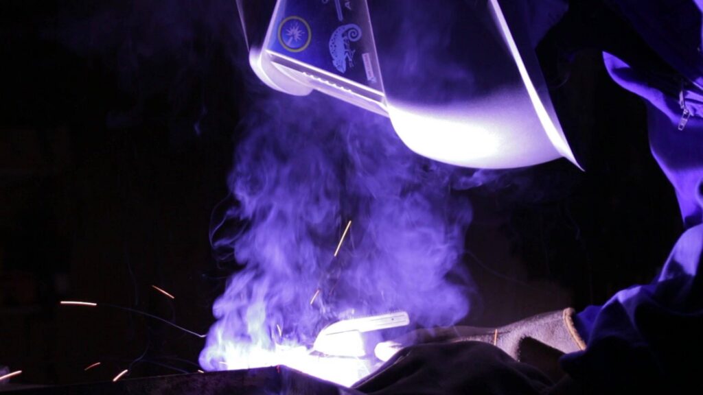 Photo de Syl portant un masque de soudure, penchée sur la soudure en cours, avec un gros arc électrique qui l'illumine, des étincelles et de la fumée.